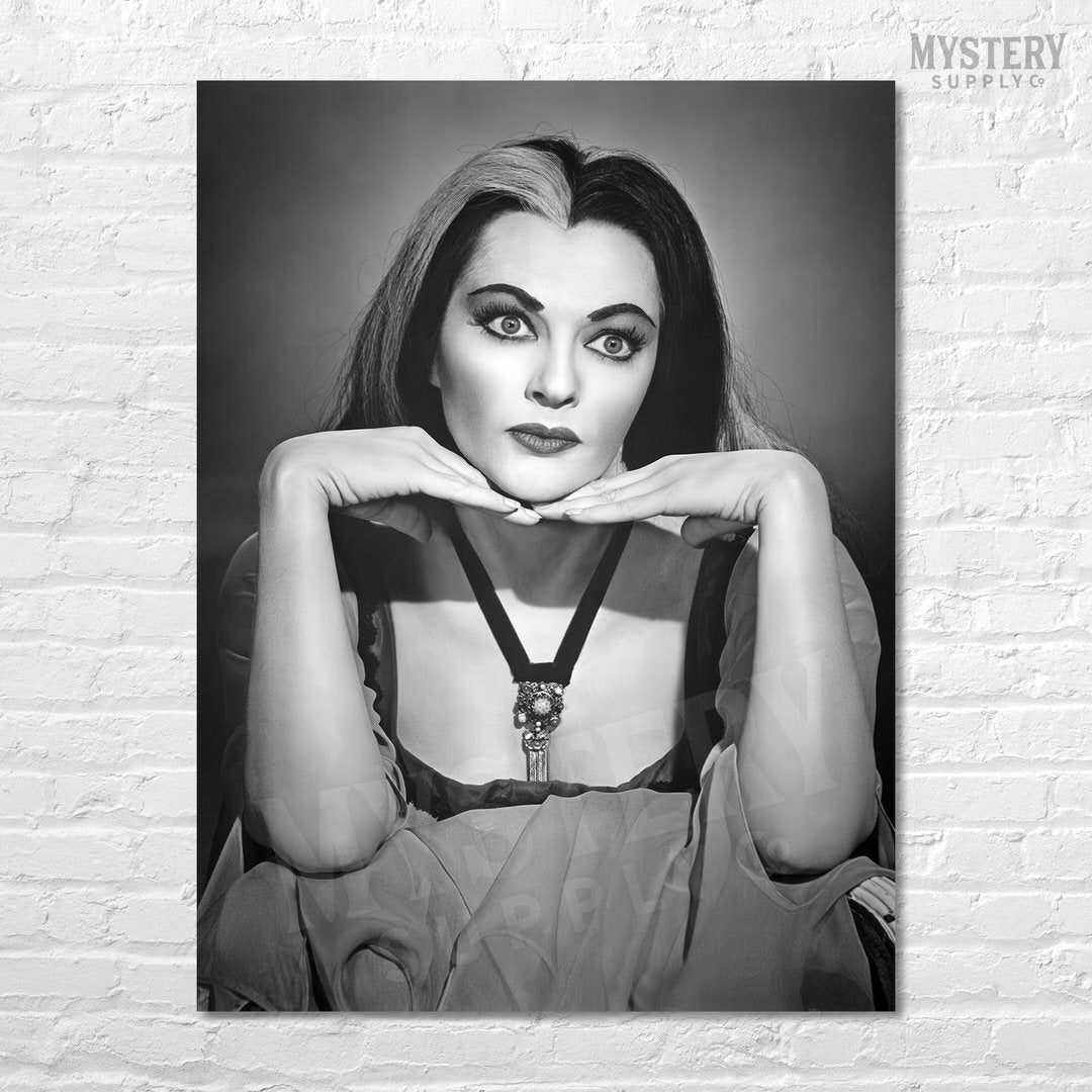 A black and white poster print of a classic film still featuring Lily Munster with dramatic makeup and parted hair, displayed on a white brick wall. A 'MYSTERY SUPPLY' watermark is visible in the top right corner.